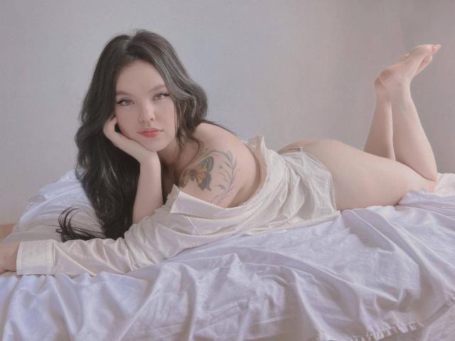 ElyMoon, laying on a bed, in a white shirt, exposing her tits and ass, with long black hair, a tattoo on her left shoulder, and legs up.