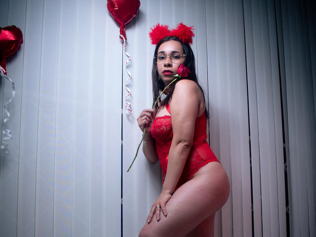 MarthaMaya with red lingerie, red furry cat ears, glasses, holding rose, red heart balloons, white vertical blinds, curvy, medium tits, pink lipstick, holding rose near her mouth.