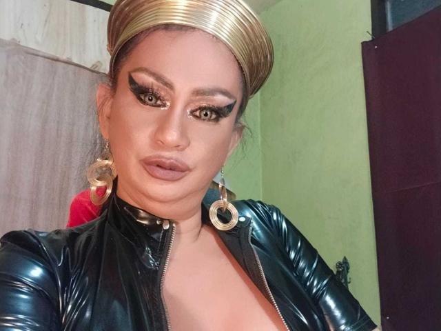 MistressKinykyJulia with golden headband, heavy eyeliner, and mascara, wearing a black shiny leather top, showing her tits, and large hoop earrings, looking at the camera.