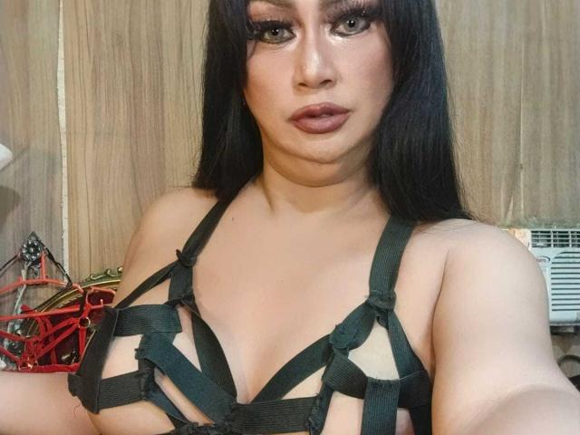 MistressKinykyJulia with long black hair, wearing a green strappy harness, exposing her tits, with full makeup and dark lipstick, in a wooden room.