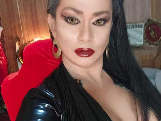 MistressKinykyJulia with glossy black hair, heavy red lipstick, gold hoop earrings, and a black leather top, showing her tits, sitting in a red chair, looking seductive.