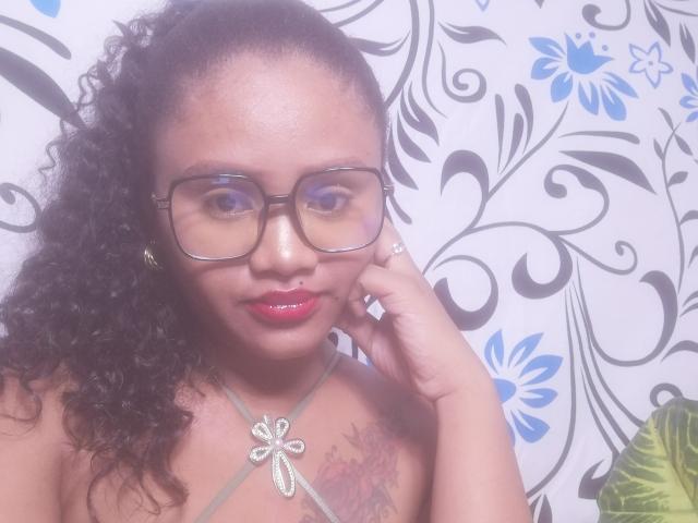 Aichamiel with curly black hair, glasses, and red lipstick, has a tattoo on her chest, wearing a necklace with a cross, and is touching her cheek.