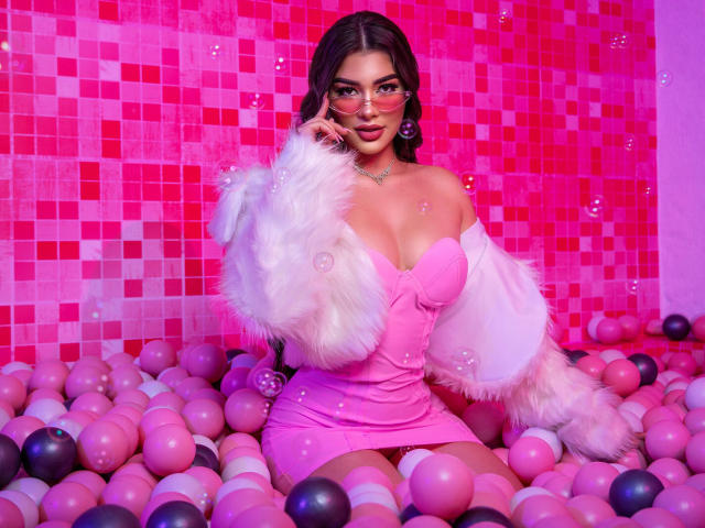SofiiaMartinez with pink dress, white fur jacket, and glasses, sitting in pink and purple ball pit, showing her tits and cute face, surrounded by pink and purple balls.