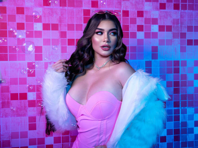 SofiiaMartinez met grote borsten, gekleed in een roze jurk zonder bandjes, een witte bontjas en een roze haarspeld, kijkt opzij, met blauwe en roze verlichting..