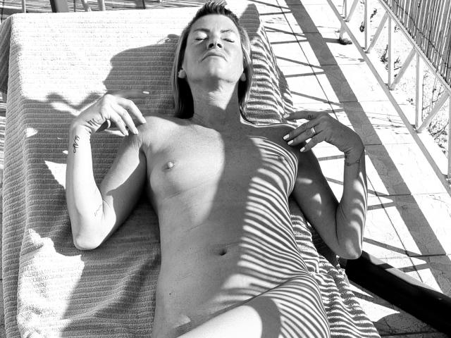 BlackAngeel, naked, lying on a sunbed with striped shadows, small tits, tattoos on right arm, eyes closed, sunbathing, hands relaxed, towel under her, outdoors, black-and-white photo.