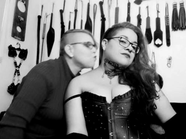 Black-and-white photo of LatinoHardGirls with glasses and dark lipstick, wearing a studded corset, and a LatinoHardBoy with a shaved head and glasses, kissing her neck.