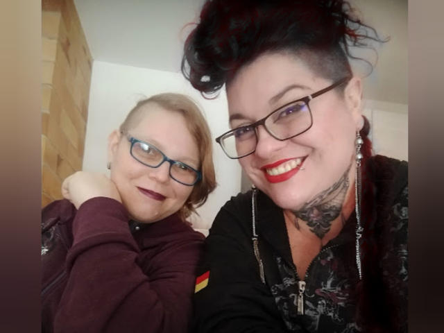Duas mulheres latinas, uma com cabelo loiro curto e óculos, a outra com cabelo escuro, batom vermelho e tatuagens, sorrindo e posando juntas. A mulher latina de cabelo escuro tem um corte tipo "undercut"..