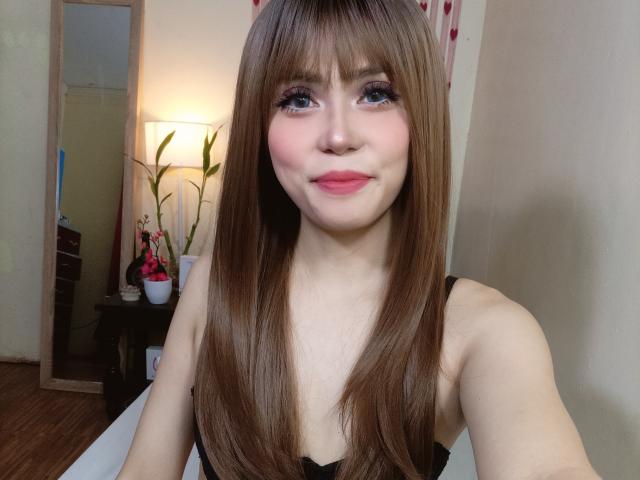 TSAnabelleSwift has long brown hair, blue eyes, pink lips, and is wearing a black top, showing her small tits and a cute smile in a bedroom selfie.