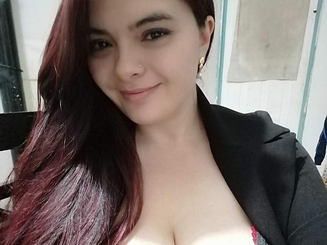 KatherinLatin with red-brown hair, smiling, wearing a black jacket, showing cleavage in a pink top, small nose ring, and gold earrings, in a selfie.
