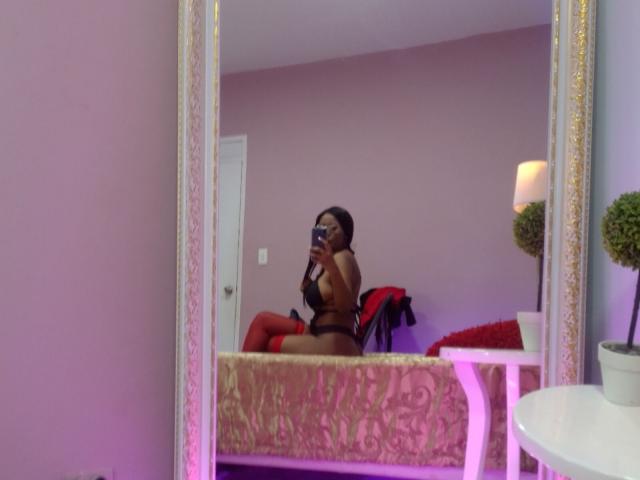 Ebonydaniels in black lingerie, holding phone, reflecting in ornate mirror, showing her tits and red thigh-highs, sitting on bed with red blanket.