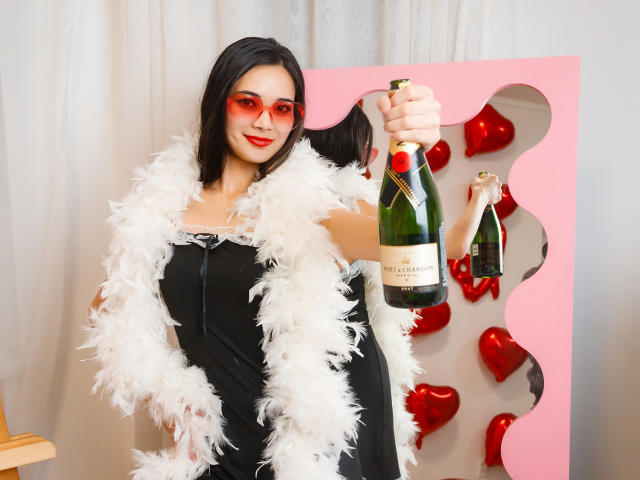 AshleyKane with red sunglasses, black dress, white feather boa, holding a bottle of Veuve Clicquot, and a mini champagne bottle, smiling, black hair, white curtain.