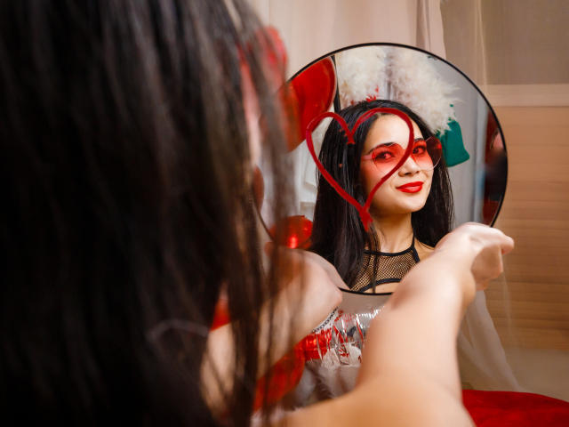Ashley Kane, con lunghi capelli neri, rossetto rosso e top in rete, che si guarda allo specchio con vernice rossa sul viso e sulle mani, in una stanza rossa..