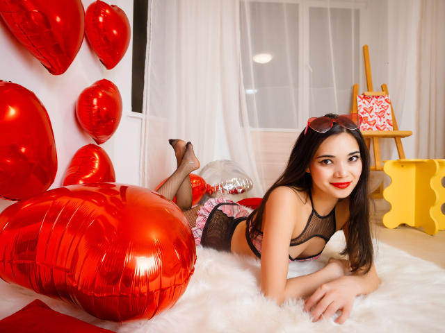 Ashley Kane con palloncini a forma di cuore rossi, sdraiata su una pelliccia bianca, che indossa lingerie in rete nera, rossetto rosso, occhiali in testa, sorridente, slip arruffati rosa e bianchi, tacchi alti rossi..
