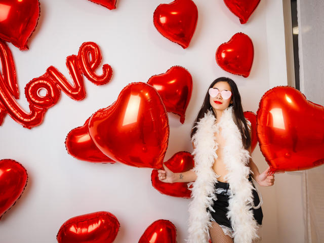 Ashley Kane con palloncini a forma di cuore rossi, che indossa una collana di piume bianche, pantaloncini neri, calze a rete e occhiali a forma di cuore rosa, tiene in mano palloncini a forma di cuore, mostra il sedere, in una foto sexy a tema San Valentino..