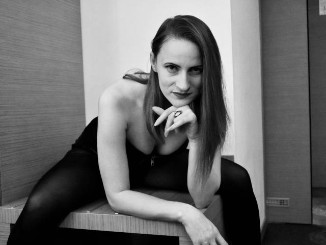 AbyX with long hair, leaning on a table, wearing a ring, black top, and black tights, showing her tits, in a black-and-white photo.