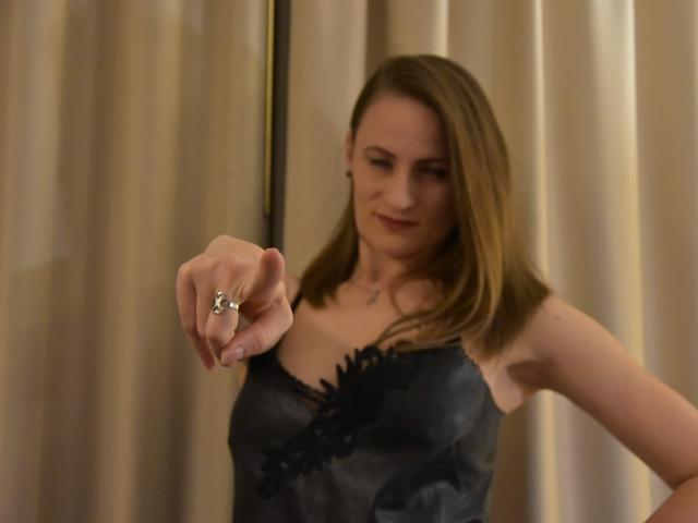 AbyX with medium brown hair, wearing a black lace-trimmed satin camisole, points at a silver ring on her index finger, showing a slight smile.