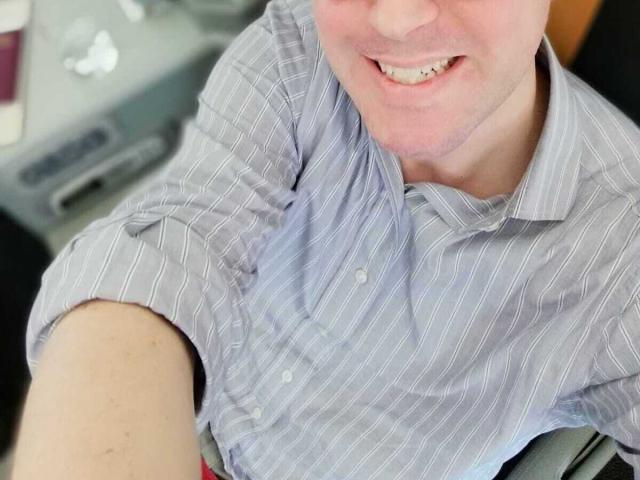 Marbosio69's face and upper body, wearing a blue striped shirt, smiling with visible teeth, head slightly tilted, left arm resting on a surface, background blurred.