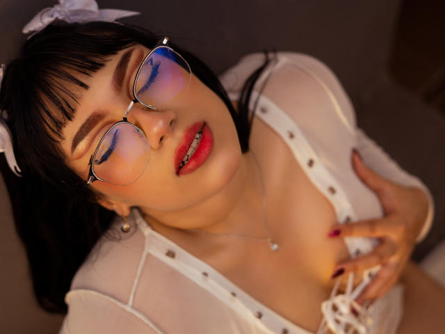 MillyJohnson with black hair, glasses, red lipstick, white sheer top, hand on tits, eyes closed, red nail polish, white hair bow, necklace, and parted lips.