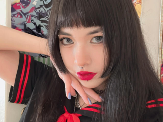 SabriPrettty with black hair, blue eyes, red lipstick, nose ring, and septum piercing, wearing a black and red sailor outfit, hand on cheek, and blue nails.