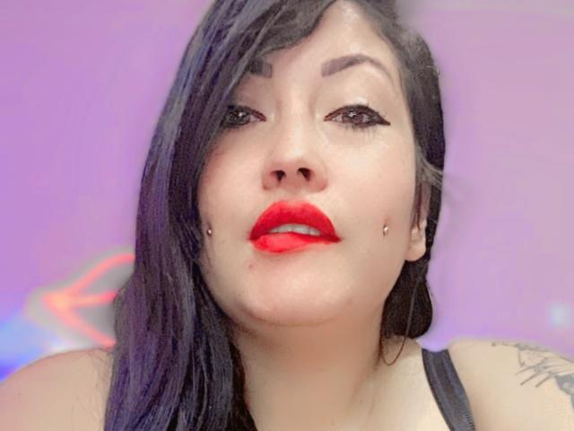 NatashaBoobsLatina has dark hair, red lipstick, black eyeliner, and two facial piercings. She's looking up with a slight pout, wearing a black tank top.