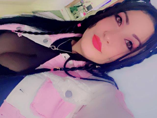 NatashaBoobsLatina con rossetto rosa, reggiseno nero e canottiera rosa e bianca, che mostra il décolleté, capelli neri intrecciati e orecchini, in un selfie..