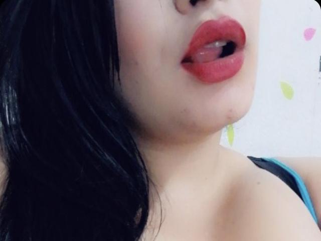NatashaBoobsLatina's face, showing her red lipstick, slightly open mouth, black hair, and fair skin, with a visible black strap and her blue top's edge.