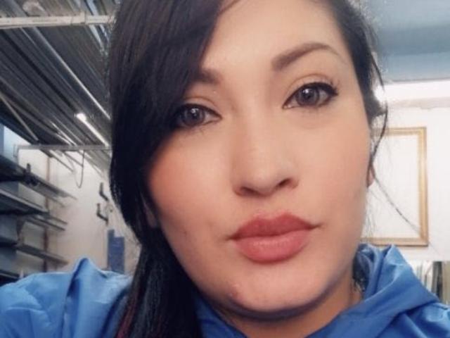 Het gezicht van NatashaBoobsLatina op een selfie, met donker haar, bruine ogen en roze lippen, gekleed in een blauw shirt, in een licht onscherpe, binnenomgeving..