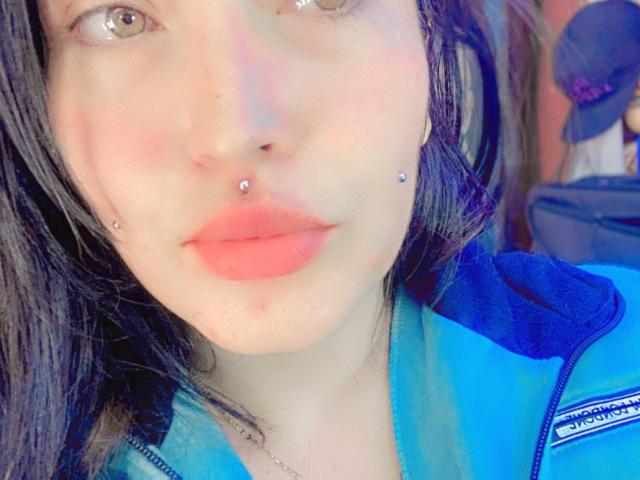 NatashaBoobsLatina met bleke huid, bruine ogen, zwart haar, neuspiercing, lipiercing, rode lippen, draagt een blauwe en zwarte jas, licht gerood, zacht licht, lichte glimlach, zachte uitdrukking..