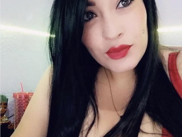 NatashaBoobsLatina, con lunghi capelli neri, rossetto rosso e un piccolo piercing sul viso, indossa una maglietta rossa, ha una collana e un'espressione sexy e sicura di sé..