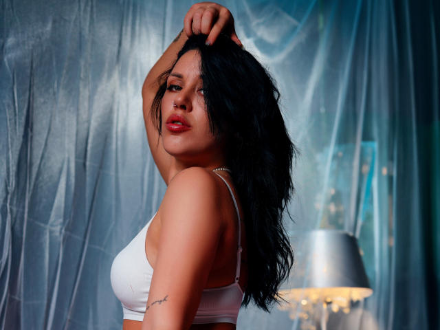 LaylaCruz with long black hair, red lipstick, white bra, raised arm, tattoo on right arm, looking at camera, soft blue lighting, silver lamp in background, sexy, confident.