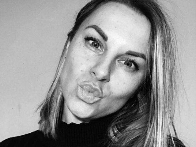 BelleLisaG's pouty face in a black-and-white selfie, her thick eyebrows and freckles stand out, wearing a turtleneck, blonde hair slightly messy, lips puckered in a kiss.