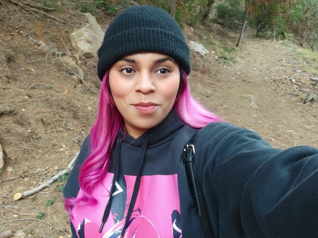 Chili with pink highlights is pouting, wearing a black beanie and hoodie with pink graphics, in a dirty forest trail.