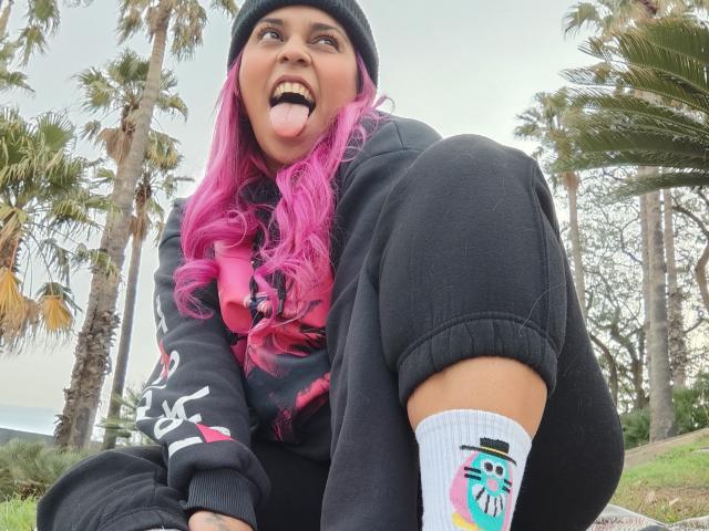 Chili, una chica punky con el pelo rosa brillante, saca la lengua mientras está en cuclillas, con una sudadera negra y calcetines con diseños de dibujos animados de colores, en un parque con palmeras.