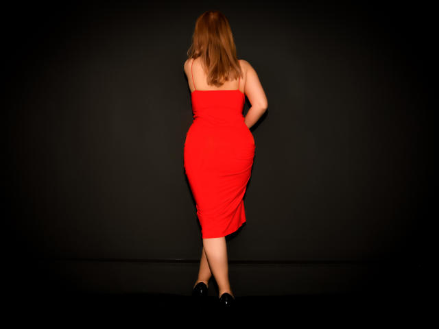 NymphoArri stands with her back to the camera, wearing a tight red dress, highlighting her curvy ass and round butt, with her hands on her lower back, in black shoes.