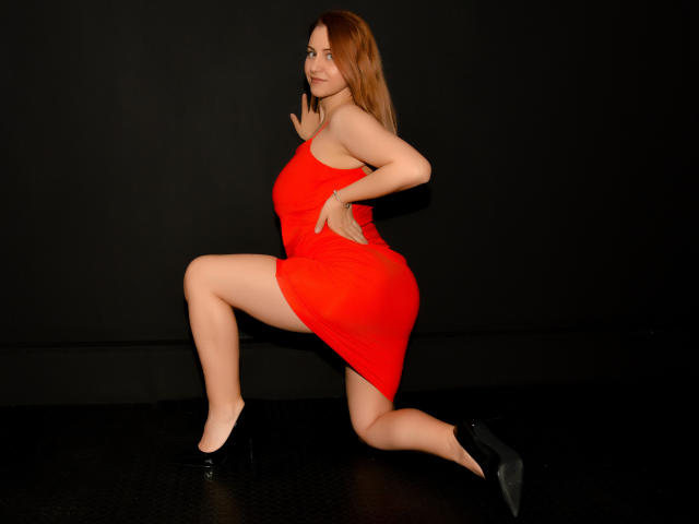 NymphoArri con un vestito rosso, inginocchiata su una gamba, che mostra il sedere e i seni, indossa tacchi neri, su uno sfondo nero. La sua mano tocca il muro..
