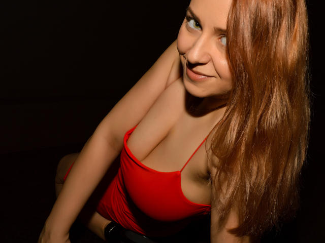 NymphoArri with red tank top, showing big boobs, smiling at camera, long brown hair, light skin, green eyes, sitting in dark, wearing black belt, looking cute and sexy.