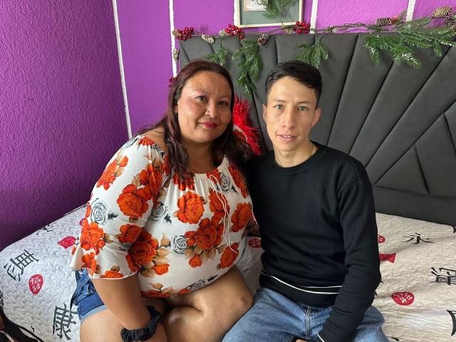 AliceAndTheo with big ass in a floral top, sitting next to Theo with a small dick in jeans, on a bed with Christmas decor. Alice has red lipstick.