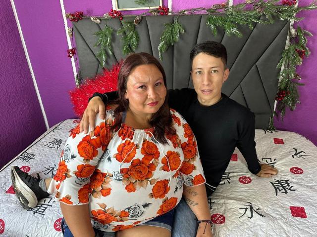 AliceAndTheo with red flowers on her shirt, big tits, and a chubby ass, sits on a bed with her arm around Theo, who has a black shirt and a cute butt.