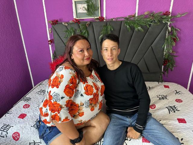 Alice und Theo, mit großen Brüsten und einem wohlgeformten Hintern, sitzen auf einem Bett. Alice trägt ein Oberteil mit Blumenmuster und Jeansshorts, während Theo ein schwarzes Langarmshirt und Jeans trägt..