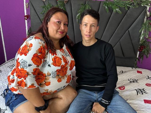 AliceAndTheo hat einen großen Hintern und trägt ein Oberteil mit Blumenmuster. Sie sitzt neben Theo, der einen kleinen Penis hat und Jeans sowie ein schwarzes Hemd trägt. Sie sitzen auf einem Bett mit roten und schwarzen Mustern..