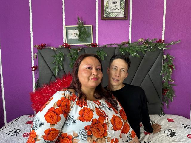 AliceAndTheo, eine mollige Frau mit roten Haaren und einem orangefarbenen Blumenkleid, sitzt auf einem Bett mit kleinen Brüsten, rotem Lametta, und ein Mann mit schwarzen Haaren in einem schwarzen Hemd lächelt..