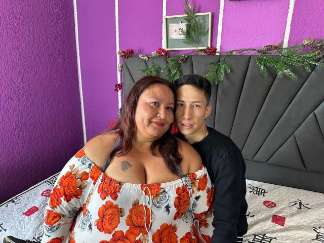 AliceAndTheo, uma mulher corpulenta com cabelo ruivo e seios grandes, vestindo um vestido floral com um ombro descoberto, está sentada em uma cama com seu namorado, que tem um corpo pequeno, veste uma camiseta preta..