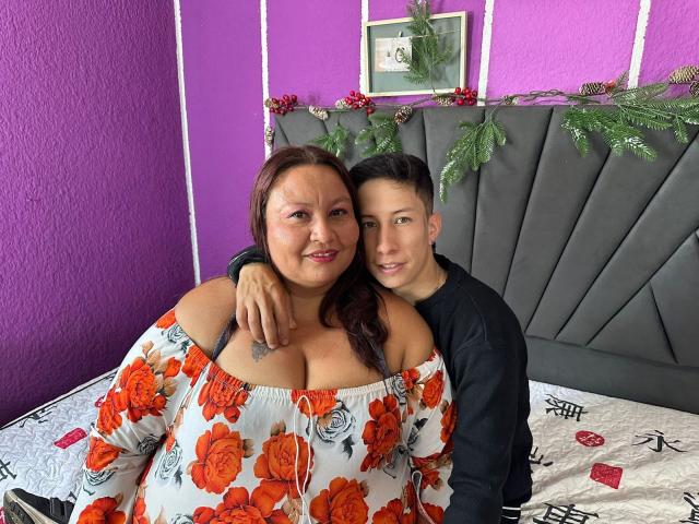 AliceAndTheo with large tits, red hair, and floral dress, sitting on bed, arm around Theo, black shirt, grey headboard, purple wall, Christmas garland, white and black bedspread.
