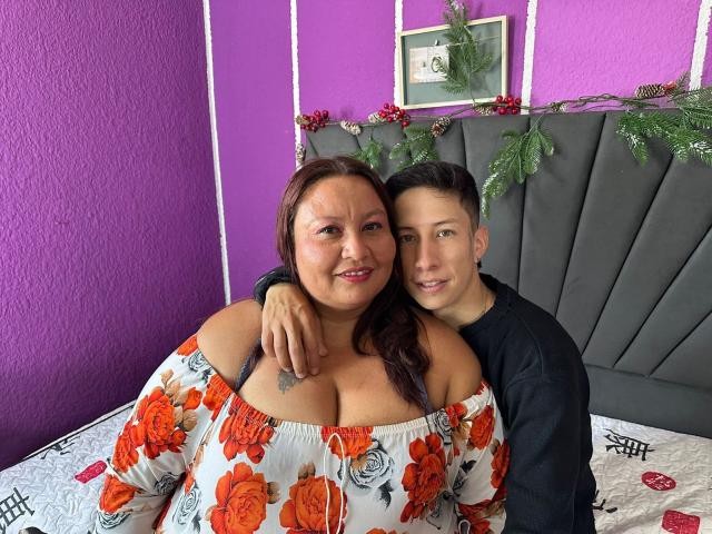 AliceAndTheo with large tits in an off-shoulder floral top and Theo with a black shirt, both smiling, Theo's arm around AliceAndTheo's shoulder, purple wall background.