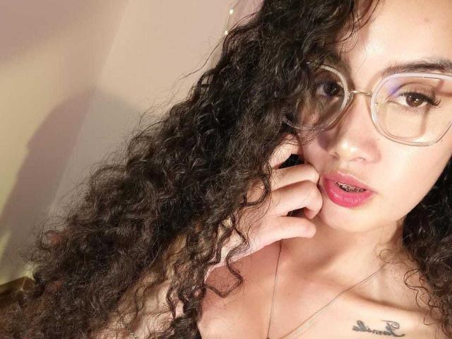 ValeriaMarie with curly black hair, glasses, pink lips, slight pout, hand near face, necklace, tattoo on chest, soft light, slightly open mouth, looking at camera.