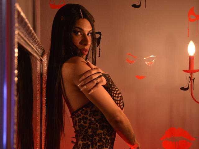 SalomeGrey with long black hair, wearing a leopard print dress and fishnet top, looking back with arms crossed, red candlelight, and red lip prints on the wall.