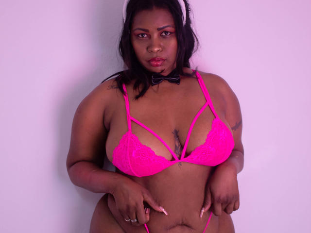 AshleyPumperHot, curvy black woman with dark skin, pink lace bra, small black bowtie, pink nails, tattoos on chest, black hair, pink lipstick, looking at camera, hands on bra.