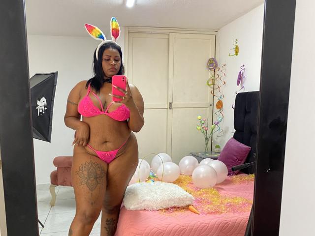 AshleyPumperHot in pink lingerie, rainbow bunny ears, holding phone, thick thighs, large ass, tattoos on left thigh, pink bra, pink panties, mirror selfie, white balloons on bed.