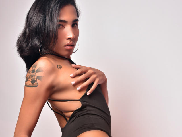 GabySantos with black hair, wearing a black top, showing a tattoo on her right shoulder, hand on her chest, hoop earrings, and a small heart tattoo above her breast.