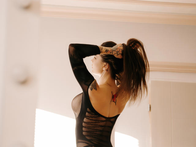 JinaMoon with arms raised, tying brown ponytail, wearing black mesh top, exposing back tattoo of red butterfly, tattoos on arms, sunlight highlighting her.